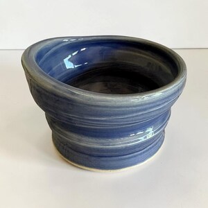 May include: A handmade ceramic bowl with a blue glaze. The bowl has a unique, ridged design around the exterior and a smooth interior. The rim is slightly irregular, adding to its handcrafted appeal. The bowl sits on a white base.