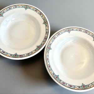 May include: Two white ceramic soup bowls with a decorative floral border in shades of red, green, and yellow. The bowls have a wide rim and a deep center, suitable for serving soups or stews. The bowls are set against a gray background.