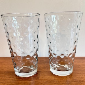 May include: Two clear drinking glasses with a textured, bubble-like pattern. The glasses are tall and cylindrical, with a slightly flared top. They are made of clear glass and sit on a wooden surface.