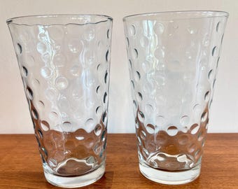 May include: Two clear drinking glasses with a textured, bubble-like pattern. The glasses are tall and cylindrical, with a slightly flared top. They are made of clear glass and sit on a wooden surface.