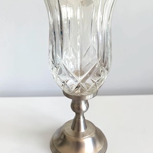 May include: A clear glass hurricane candle holder featuring a diamond-cut geometric pattern on the upper bowl. The glass sits atop a brushed silver-toned metal pedestal base with a circular foot and a smooth, tiered stem design.
