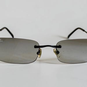 May include: Rimless sunglasses with rectangular grey-tinted lenses and thin black metal temples. The left lens features the white Ray-Ban logo in the upper corner. The frame includes adjustable nose pads and a thin black bridge connecting the lenses.
