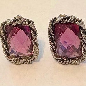 May include: A pair of silver-toned stud earrings. Each earring features a rectangular, faceted, magenta-colored stone set within a twisted rope-like border. The earrings are designed for pierced ears and are displayed against a neutral background.