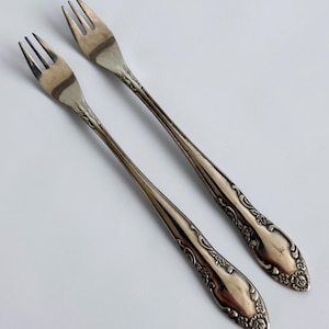May include: Two silver-colored forks with three tines each. The handles are decorated with floral patterns. The forks are angled on a white background, showcasing their design and detail.