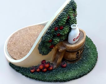 Op de afbeelding: Een decoratieve onderzetterhouder met een gebeeldhouwde appelboom met rode vruchten, een houten vat en een witte kan met het opschrift CIDER. De basis is groen getextureerd als gras en de binnenruimte is voorzien van een bruine kurken inzet.
