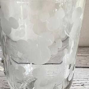 Pu&ograve; includere: Tazza di vetro trasparente con base arrotondata e manico. La tazza &egrave; decorata con un motivo di disegni bianchi a forma di Mickey Mouse. Il vetro ha un leggero aspetto smerigliato. La tazza &egrave; su una superficie di legno.