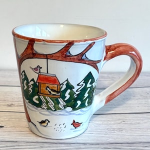 May include: White ceramic mug with a winter scene featuring a bird feeder, evergreen trees, and birds. The handle and rim are painted in a reddish-brown color. The mug has a wide, slightly flared shape.