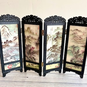 May include: A four-panel folding screen with a black frame and ornate top. Each panel features a landscape painting with mountains, trees, and water in muted colors. The screen is freestanding and decorative.