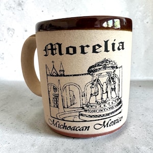 May include: Tan ceramic mug with a brown rim and handle. The mug features a black illustration of a building and fountain, with the word "Morelia" and "Michoacan Mexico" printed on the front. The mug is a standard size.