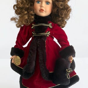May include: A porcelain doll with curly brown hair and blue eyes, wearing a red velvet coat trimmed with black faux fur and gold accents. The doll has a black hat and matching muff. A gold tag reads "Edition Limitee".