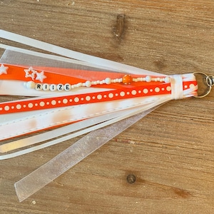 May include: A handmade keychain with orange and white ribbons, clear plastic strips, and a beaded strand spelling "RIIZE". The keychain includes star-shaped embellishments and a metal clasp.