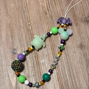 May include: A Halloween-themed beaded charm with a white string loop. The charm features a purple bat, a glowing green ghost, and a mouse-shaped bead. The word "BOO" is spelled out with letter beads, and the charm includes various colorful beads.