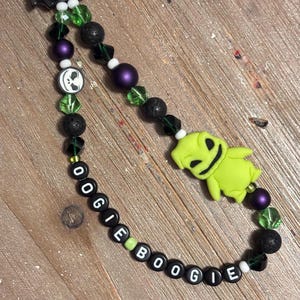 May include: A handmade beaded lanyard featuring a black bat charm, green and purple beads, and a lime green Oogie Boogie figure. The lanyard spells out "OOGIE BOOGIE" with black and white letter beads. The lanyard is designed with a variety of bead shapes and textures.