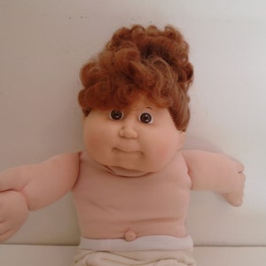 May include: A Cabbage Patch Kid doll with brown curly hair, brown eyes, and a pink and white outfit.