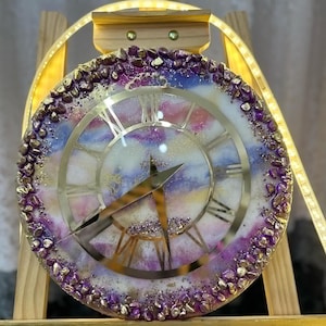 May include: A round clock with Roman numerals and a white face with purple and gold accents. The clock is surrounded by purple and gold glitter and is displayed on a wooden easel.