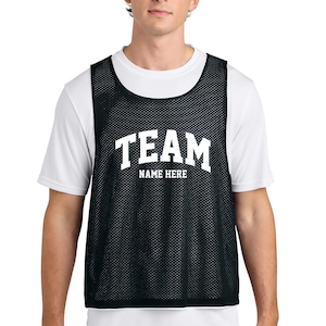May include: A black mesh athletic pinnie worn over a white short-sleeved t-shirt. The black pinnie features the white text TEAM and NAME HERE printed in a bold, block-style font across the center of the chest area.