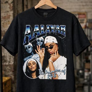 May include: Black t-shirt featuring a graphic of Aaliyah with the text "AALIYAH" in a blue and white design. The shirt includes the text "The Princess of R&B" below the image of Aaliyah.