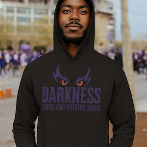 May include: Black hooded sweatshirt with the word "DARKNESS" in purple, above the phrase "THERE AND NOTHING MORE". The design includes purple and red stylized eyes. The person is wearing the hoodie.