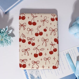 May include: A rectangular cream-coloured case with a repeating pattern of red cherries and bows. The design includes small diamond shapes. The case is likely for a phone or similar device.