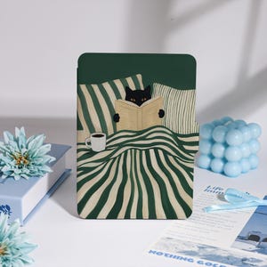 May include: A green tablet case with a black cat reading a book in bed, accompanied by a white coffee mug. The design incorporates green and white striped bedding and pillows. A light blue bubble cube and a blue flower are also present.