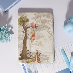 May include: A rectangular e-reader cover featuring a printed illustration of Winnie the Pooh characters including Piglet, Eeyore, and Tigger in a forest setting. Pooh is shown holding a red balloon while hanging from a tree branch in the scene.