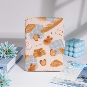 May include: A light-colored case featuring a whimsical bakery-themed design. The case is adorned with illustrations of croissants, bread, and bunny-shaped pastries. The text "cong baker" is visible on the case.