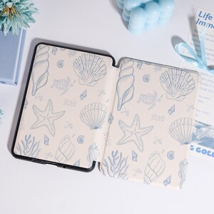Puede incluir: Funda de tableta negra abierta con interior crema y un patr&oacute;n de conchas marinas azules. El dise&ntilde;o incluye estrellas de mar, conchas y coral. La funda est&aacute; sobre una superficie blanca con otros elementos decorativos azules.