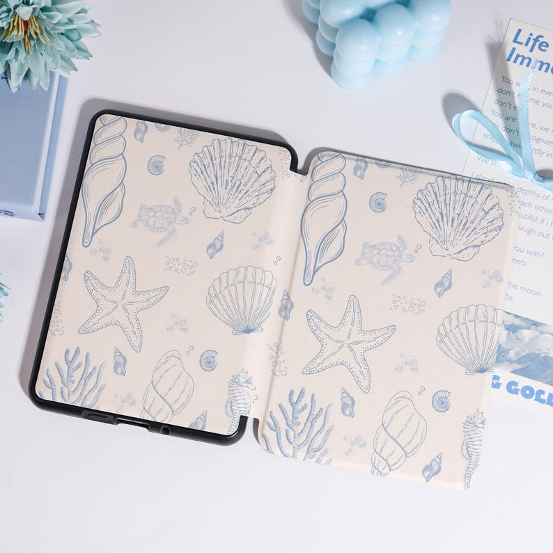 Puede incluir: Funda de tableta negra abierta con interior crema y un patr&oacute;n de conchas marinas azules. El dise&ntilde;o incluye estrellas de mar, conchas y coral. La funda est&aacute; sobre una superficie blanca con otros elementos decorativos azules.