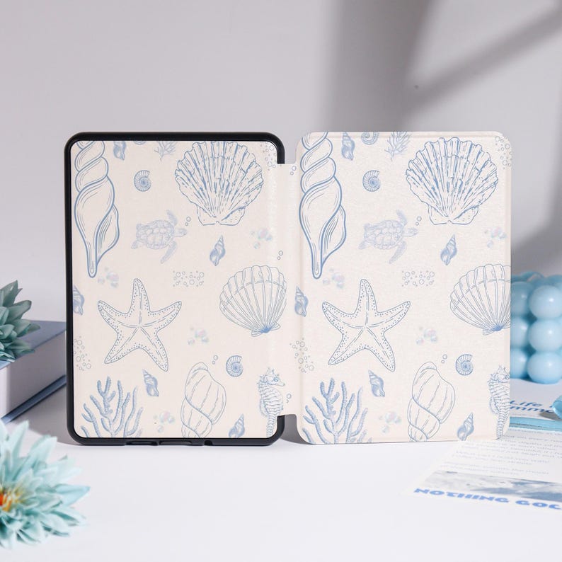 Puede incluir: Una funda de tableta color crema con un patr&oacute;n de conchas marinas azules. El dise&ntilde;o incluye estrellas de mar, conchas, caballitos de mar y coral. La funda est&aacute; abierta, revelando el interior con el mismo patr&oacute;n. La funda tiene un borde negro.