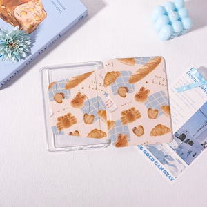 May include: A clear plastic card holder with a patterned insert featuring baked goods and bunny illustrations. The design includes croissants, bread, and the text "cong baker" on a light blue checkered background.