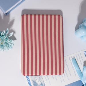 May include: A rectangular case featuring a vertical striped design in alternating red and pale pink. The case is set on a white surface, with a blue book and a light blue flower nearby.