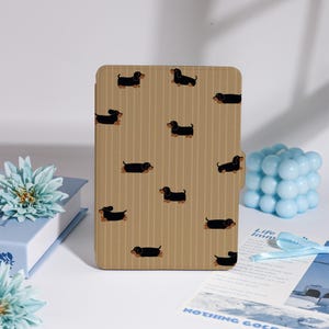 Peut inclure: Étui de tablette beige avec un motif répété de chiens teckels noirs. L'étui présente des rayures verticales et une fermeture magnétique. Le design est ludique et convient aux amoureux des chiens. L'étui est rectangulaire.