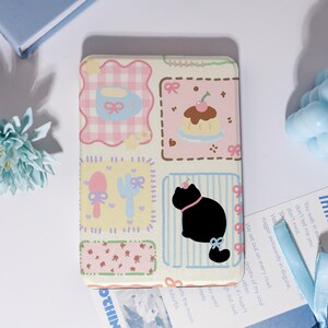 May include: A rectangular phone case with a patchwork design. The case features squares with a teacup, cake, utensils, a cat, and floral patterns. Colors include pink, yellow, and blue, with a cream-colored background. The cat has a pink collar and bow.