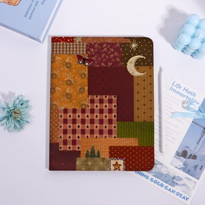 May include: A rectangular notebook featuring a patchwork-style cover design with various patterns including brown plaid, dark red stars, gold floral motifs, a crescent moon, a butterfly, and small evergreen trees in a warm, autumnal color palette.