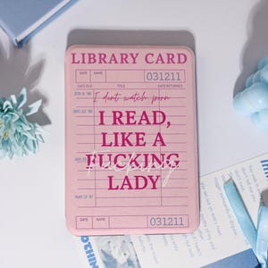 May include: A rectangular pink e-reader case featuring a vintage library card design printed in dark pink. The text reads LIBRARY CARD, 031211, I dont watch porn, I READ, LIKE A FUCKING LADY, and includes columns for date, name, and date returned.