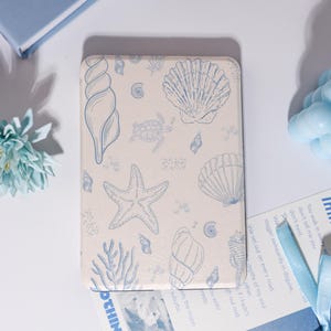 Puede incluir: Una funda rectangular blanquecina con un dise&ntilde;o de conchas marinas y estrellas de mar azules. La funda tiene un borde redondeado y est&aacute; decorada con varias criaturas marinas. El dise&ntilde;o es de color azul claro.