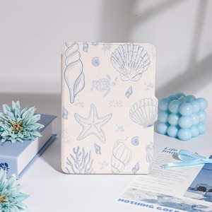 May include: A cream-colored tablet case featuring a blue seashell and starfish pattern. The case has a magnetic closure. A blue bubble candle and a blue flower are also visible.
