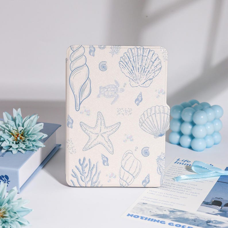 Puede incluir: Una funda de tableta color crema con un dise&ntilde;o de conchas marinas y estrellas de mar azules. La funda tiene un cierre magn&eacute;tico. Hay otros elementos decorativos azules presentes, como una vela y un libro.