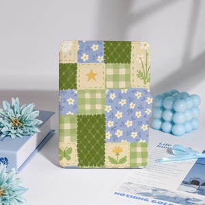 Op de afbeelding: Een vierkante notitieboekcover in patchwork-stijl met verschillende patronen, waaronder ruitjes, bloemen en effen groene vierkanten. De cover heeft een gele ster, een vlinder en een bloem. Het kleurenpalet omvat blauw, groen en crème.