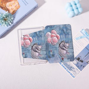 May include: A clear plastic card holder with a blue and white design featuring Eeyore holding pink balloons. The background has text that reads "I NEED YOU". A blue book and a blue candle are in the background.