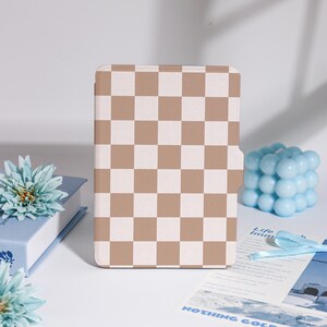 May include: A tablet case with a tan and white chequered design. The case is rectangular and has a magnetic closure. The background includes a light blue cube candle, a blue book, and a blue flower.