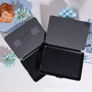 Puede incluir: Un juego de tres fundas de tableta grises oscuras abiertas. Una funda tiene una correa el&aacute;stica gris. El interior de una funda tiene una superficie negra texturizada. Las fundas est&aacute;n sobre una superficie blanca con un libro y decoraciones azules en el fondo.