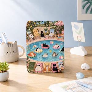 May include: A rectangular metal tin featuring a colorful illustration of various cats relaxing by and swimming in a blue pool. The scene includes cats on lounge chairs, floating on inflatable rings, and wearing sunglasses, with text on a stack of books.