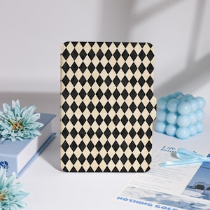 May include: A rectangular case with a black and cream diamond pattern. The case has a magnetic closure. A blue floral decoration and a blue cube-shaped candle are in the background.