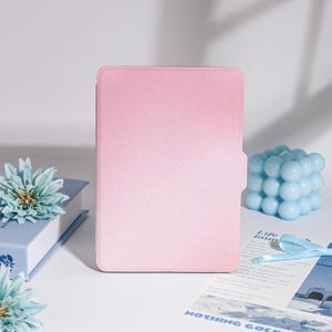 Puede incluir: Una funda de tableta rosa degradado se alza, con una vela de cubo de burbujas azul claro y una flor azul a la izquierda. También se ven un libro azul y un trozo de papel con texto. La funda tiene una textura suave.