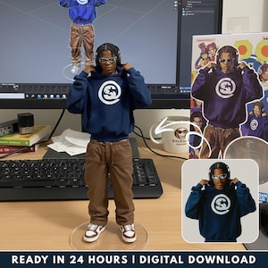 May include: A figurine of a person wearing a navy blue hoodie with a white logo, brown pants, and white sneakers with brown accents. The figurine is on a clear stand. The image also includes a computer screen and a mug.
