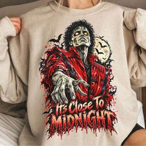 May include: A beige crewneck sweatshirt featuring a graphic of a zombie-like figure in a red jacket with glowing yellow eyes. The design includes bats, a full moon, and the text "It's Close To MIDNIGHT" in a dripping, blood-red font style.