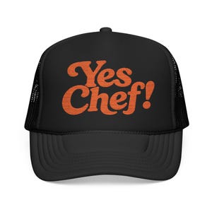 May include: A black trucker hat featuring a mesh back and a solid front panel. The front displays the text Yes Chef! embroidered in a bold, orange, retro-style font. The cap includes a curved brim and a structured crown design for a classic look.