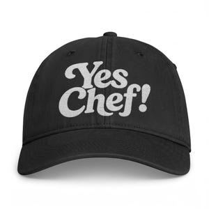 May include: A black baseball cap featuring the white embroidered text Yes Chef! in a bold, stylized font across the front panels. The cap has a curved brim, a structured crown with ventilation eyelets, and a button at the top center.