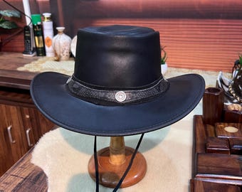 May include: A black leather cowboy hat with a wide brim and a decorative band. The hat is displayed on a wooden stand and has a chin strap. The hat appears to be approximately 14 inches in diameter.